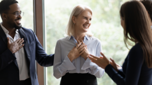 A professional woman with a warm smile places her hands on her chest while receiving compliments from her colleagues in a bright office setting. The group appears engaged and appreciative, reflecting themes of self-worth, recognition, and the challenge of accepting praise.