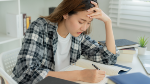 A stressed teenage girl wearing a plaid shirt sits at a desk, holding her forehead while writing in a notebook. She appears overwhelmed as she studies, surrounded by textbooks and papers. The image reflects the anxiety and fear of forgetting something often experienced by neurodiverse students.