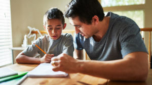 A father and his young son sit at a wooden table, working together on homework. The boy, wearing a gray t-shirt, holds a pencil while looking at his notebook with focus. The father, also in a gray t-shirt, leans in, guiding him through the task. Natural light streams through the window, creating a warm and supportive atmosphere.