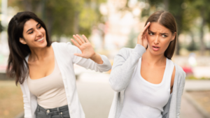 A smiling woman enthusiastically waves at another woman, who looks visibly uncomfortable and overwhelmed. The second woman places a hand on her temple, appearing frustrated or drained. This image represents the exhaustion of forced positivity and the emotional disconnect that comes from pretending to be okay.