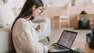 A woman wearing glasses and a cozy sweater sits on the floor with a laptop on her lap and a coffee cup in hand. She appears focused as she works in a warm, modern living space with neutral tones.