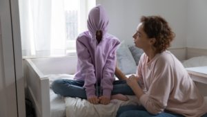 Mother sits on a bed next to her child, who is wearing a hoodie pulled tightly over their face, avoiding eye contact. The mother looks concerned, gently placing a hand on the child's arm, reflecting emotional distance and withdrawal.