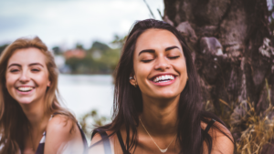 Two young women laughing outdoors, radiating joy and relaxation. Their carefree expressions contrast with the internal struggles many professionals with anxiety face—finding contentment in the present rather than constantly chasing the next goal.