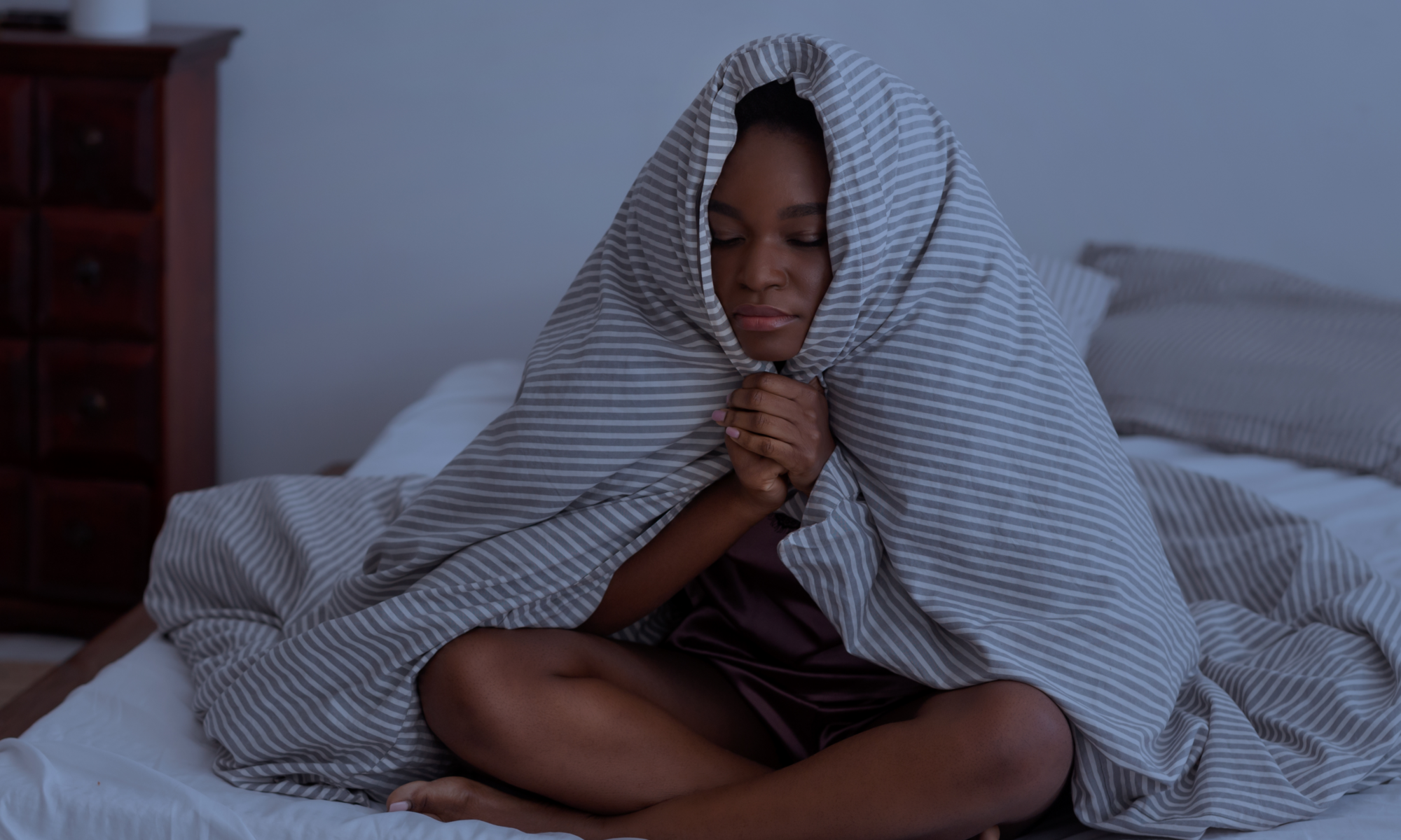 A young girl sitting on her bed at night, wrapped in a striped blanket with her eyes closed, looking restless and unable to sleep. The dim lighting and her expression convey frustration and difficulty winding down.
