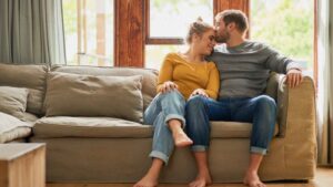 Couple sitting closely on a couch, the man gently kissing the woman’s forehead as she leans into him with her eyes closed, symbolizing comfort, presence, and the quiet intimacy of slowing down and savoring the moment.