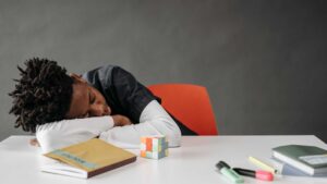Tired child resting their head on a desk, surrounded by school supplies and a puzzle cube, suggesting exhaustion and difficulty focusing, possibly due to lack of sleep.