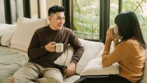 A man and a woman sit on a couch with mugs in hand, engaged in quiet conversation. The setting is calm and intimate, with natural light streaming in through large windows overlooking green foliage. The man appears to be speaking while the woman listens, evoking a sense of trust, connection, and emotional vulnerability.