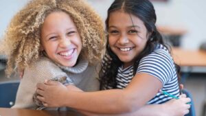 Two young girls smiling and hugging each other in a classroom setting. They appear happy and connected, suggesting themes of friendship, support, and emotional warmth.