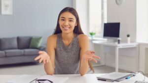 Smiling young woman sitting at a desk with open hands, as if in conversation or explaining something, in a bright modern home office with a laptop, tablet, and glasses in front of her.