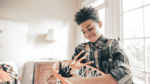A young boy smiles gently while playing with a small basketball hoop indoors. His expression is calm and focused, capturing a quiet moment of joy. The image reflects the theme of overgiving and internal pressure to be ‘good,’ central to the article on neurodiverse children struggling with guilt, boundaries, and self-worth.