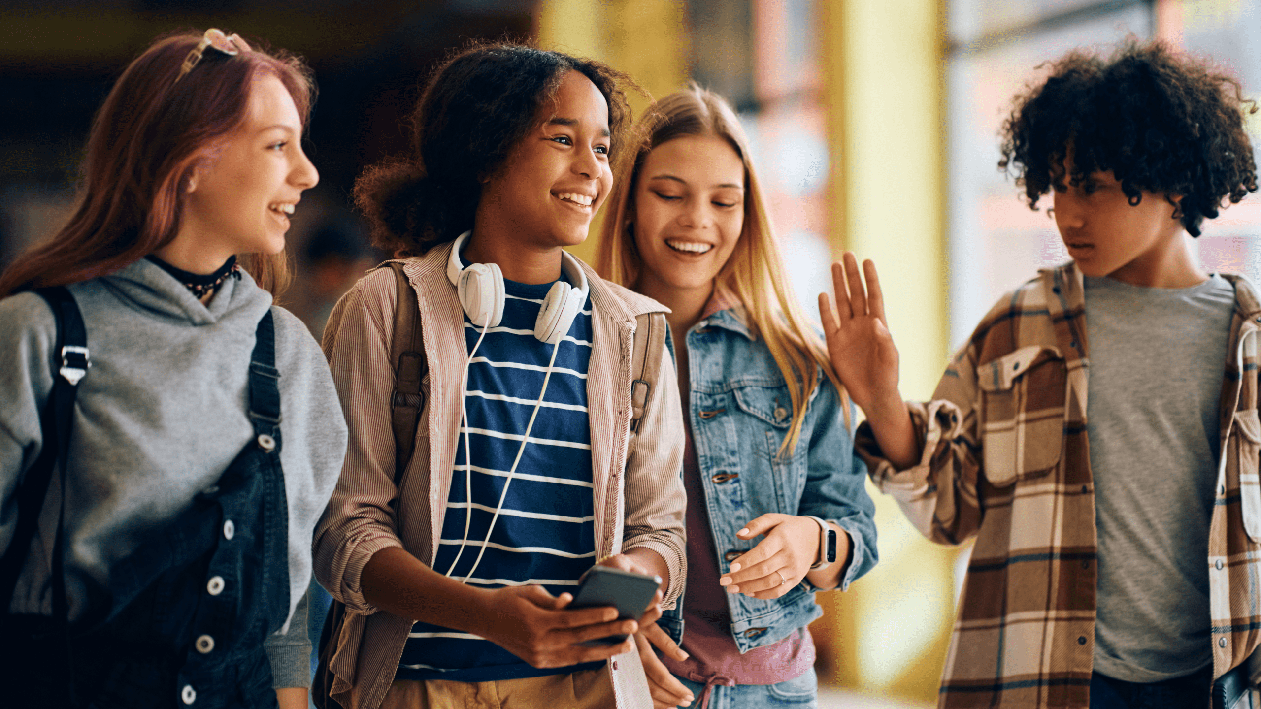A group of teenagers walks through a school hallway, smiling and chatting. One girl, wearing headphones around her neck and holding a phone, is at the center of the group, laughing along with her friends. While she appears cheerful, the image reflects the hidden emotional toll of always being the ‘easy-going one’—masking exhaustion and anxiety to maintain social harmony.