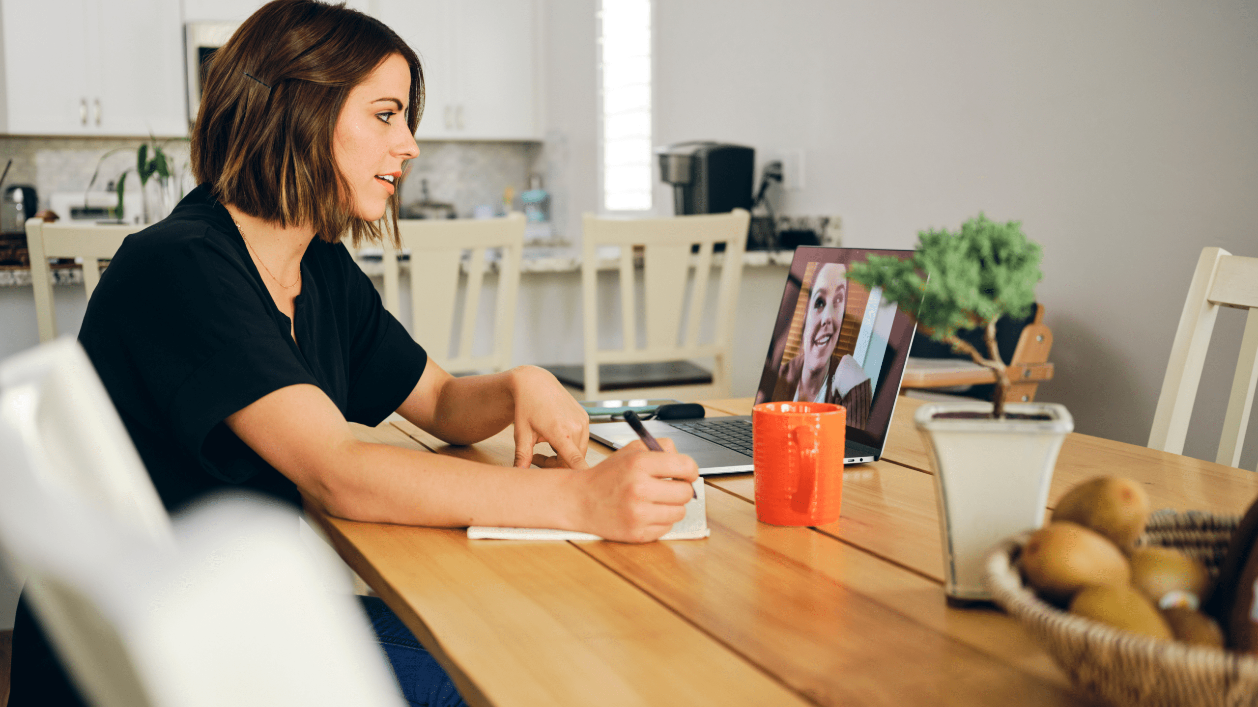A woman sits at a wooden dining table, engaged in a video call on her laptop. She is writing in a notebook while listening attentively. The setting is a cozy, well-lit home kitchen with a plant, an orange coffee mug, and a basket of fruit on the table, creating a warm and focused atmosphere.