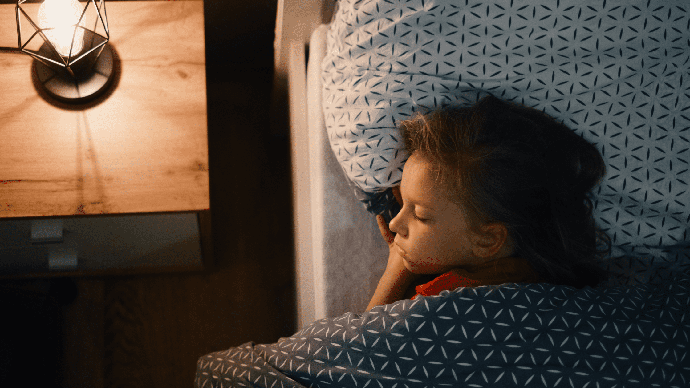 A young child sleeping peacefully in bed under a patterned duvet, with a warm bedside lamp glowing on a wooden nightstand.