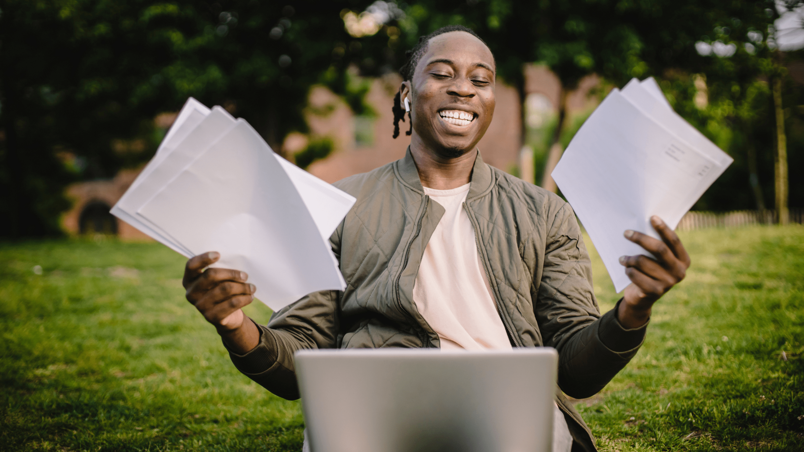 A young man sits outdoors on a grassy field with a laptop in front of him, smiling brightly while holding multiple sheets of paper in both hands. He appears joyful and accomplished, possibly celebrating a moment of success or progress.