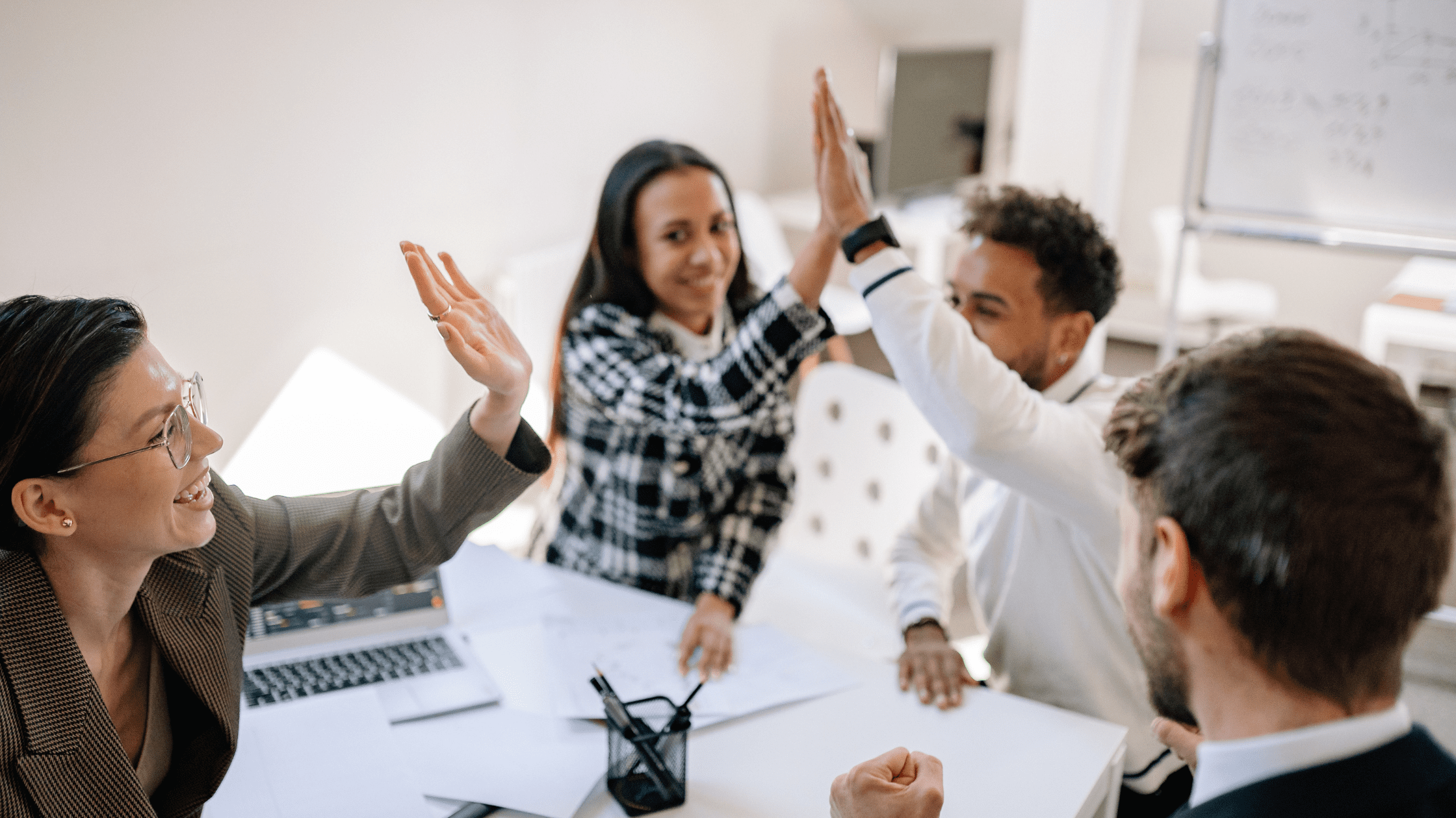 Four professionals smiling and giving high-fives around a desk in a bright office, celebrating teamwork and success.