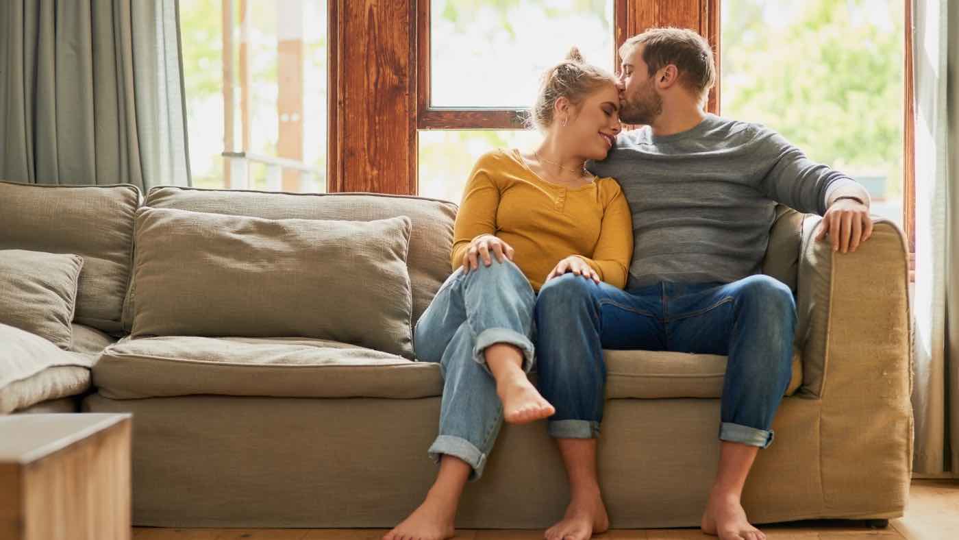 Couple sitting closely on a couch, the man gently kissing the woman’s forehead as she leans into him with her eyes closed, symbolizing comfort, presence, and the quiet intimacy of slowing down and savoring the moment.