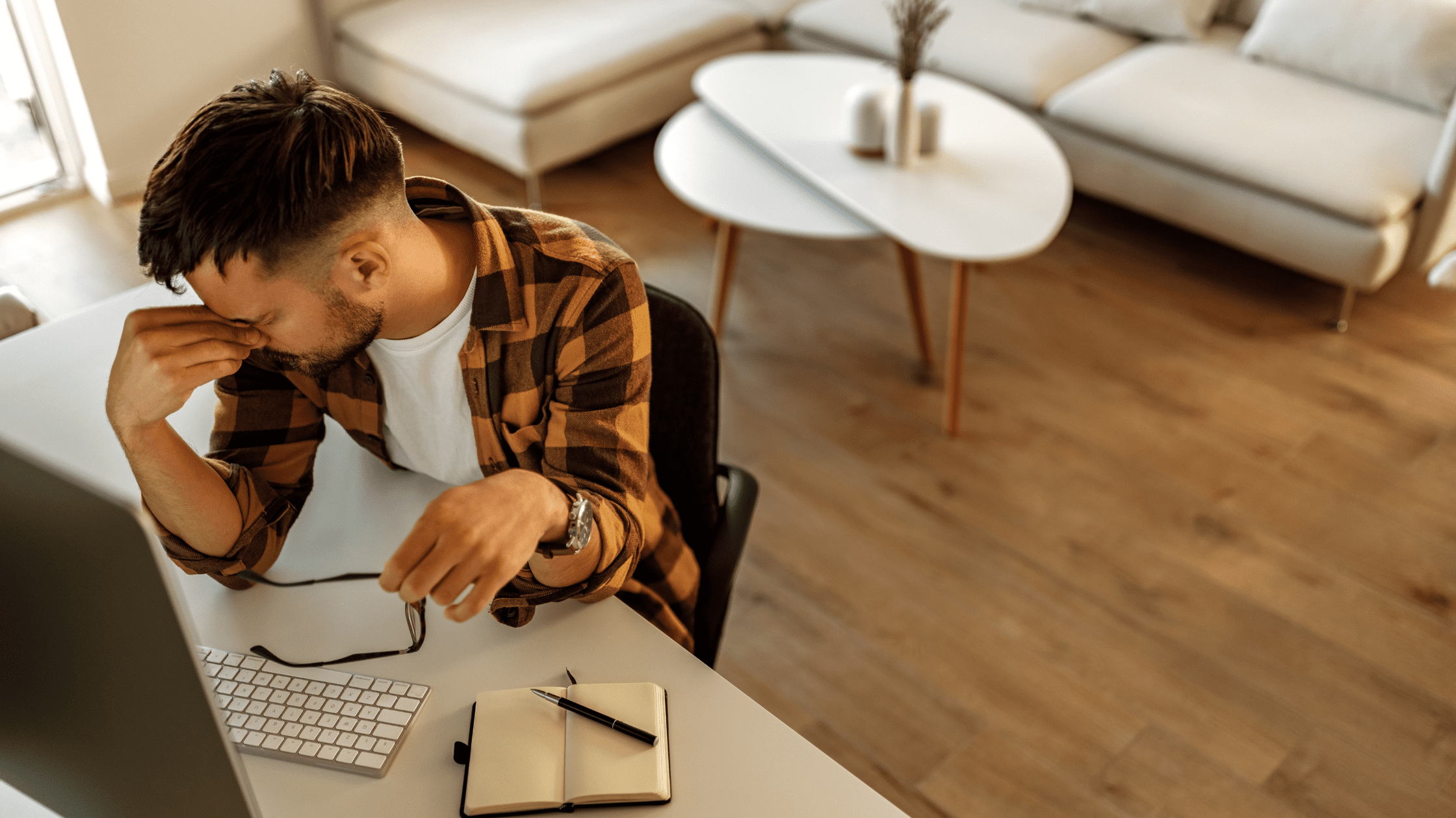 Stressed young professional sitting at a desk, holding glasses and rubbing eyes in frustration while working from home.