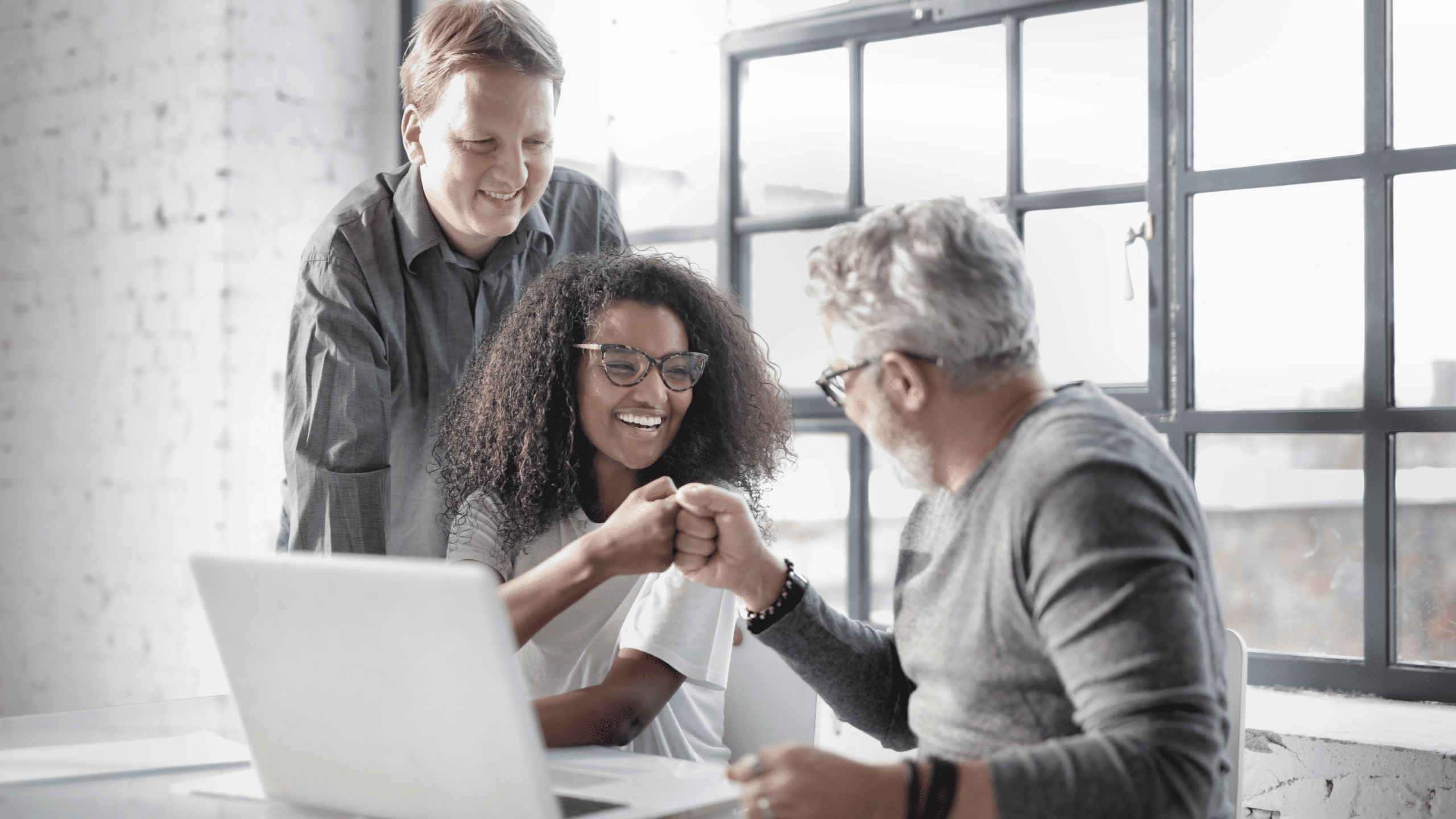 Three diverse colleagues in a bright office space celebrating teamwork with smiles and a fist bump, conveying positivity and collaboration.