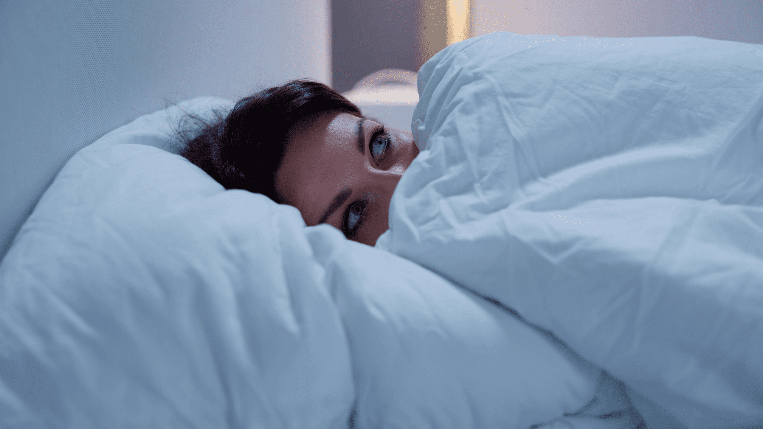 A woman lying in bed, partially covered by a white blanket, with wide-open eyes staring into the distance. The dim lighting suggests it’s nighttime, and she appears restless and unable to sleep.
