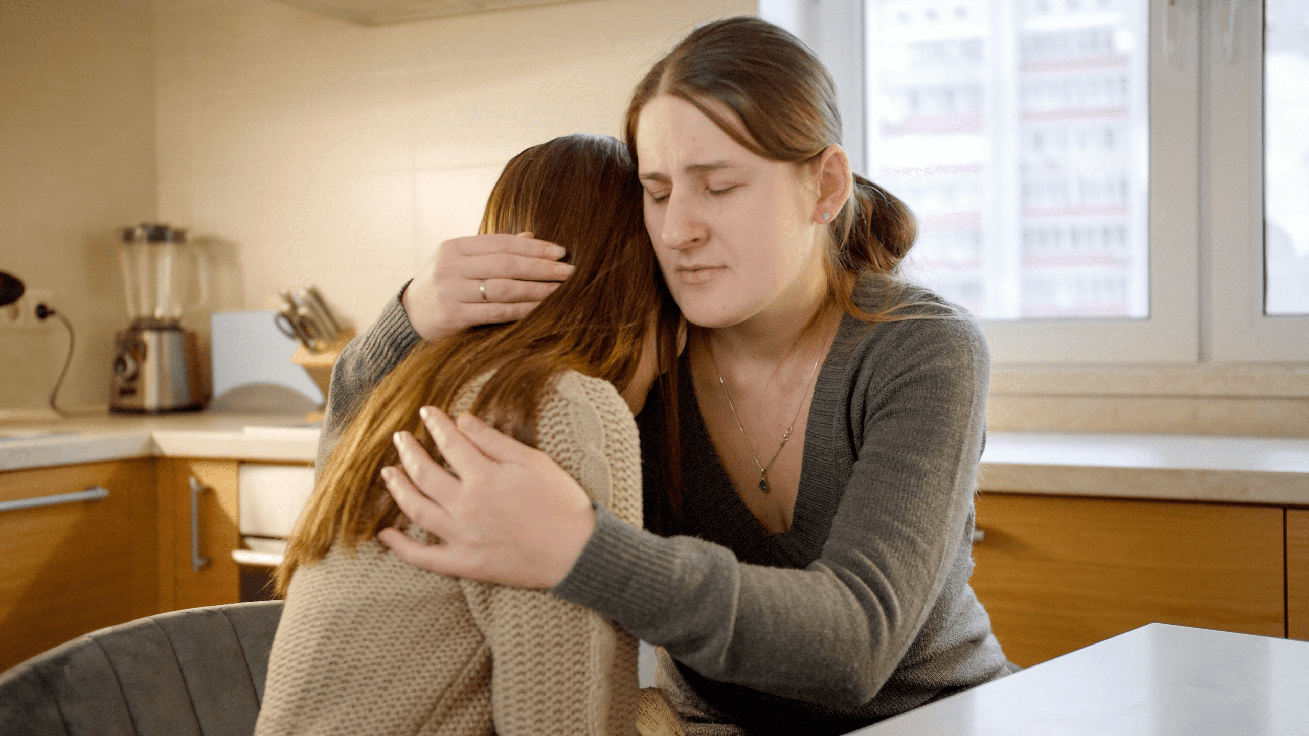 A mother sits beside her daughter, gently holding her. The daughter leans into her mother’s embrace, seeking comfort and reassurance. Soft lighting highlights their emotional connection in a moment of support and understanding.