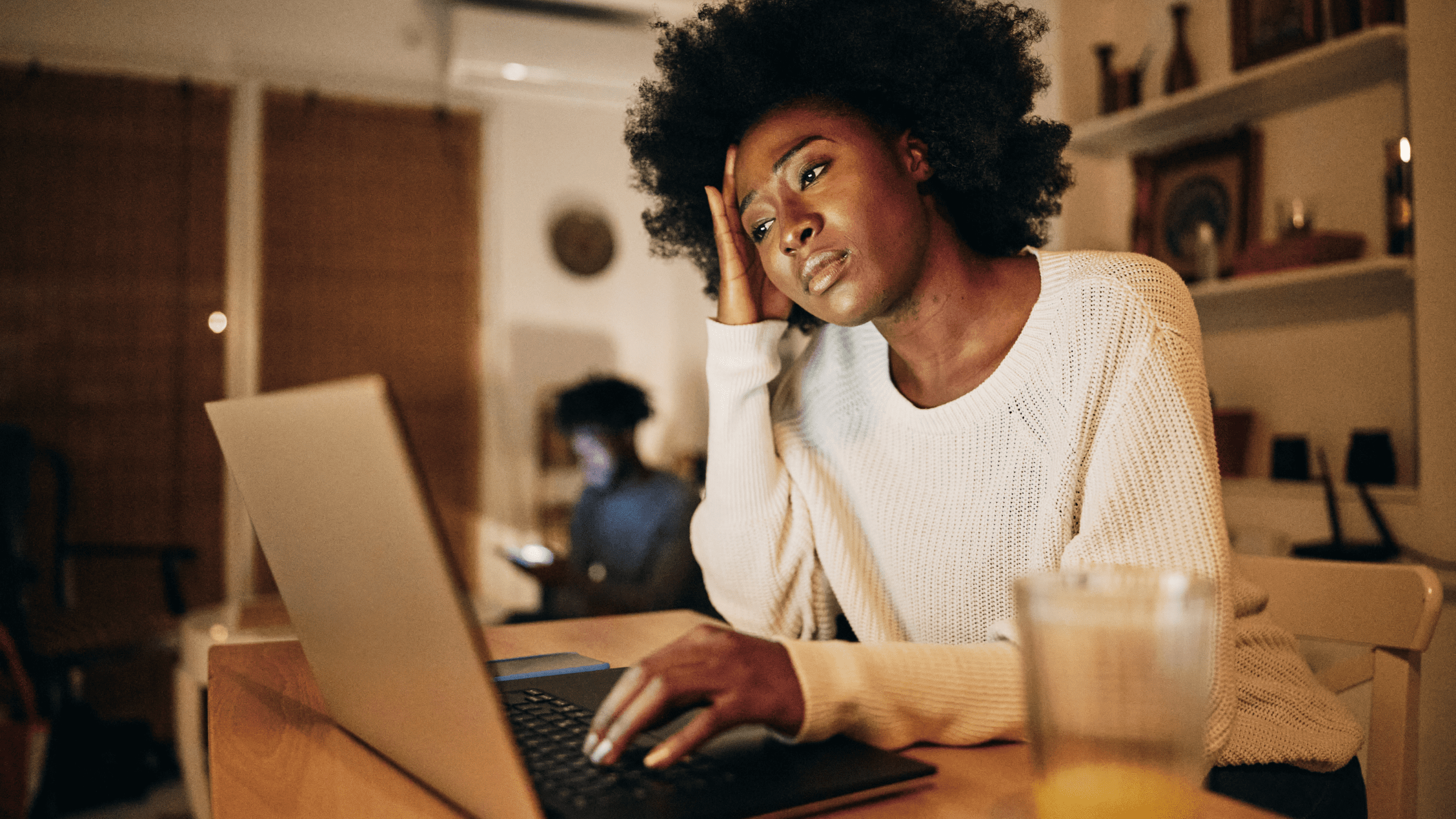 A young woman with natural curly hair sits at a wooden table, looking frustrated as she rests her head on her hand while working on a laptop. A dimly lit room with shelves and a person using a phone in the background suggests a late-night setting. A half-empty glass of juice sits nearby, emphasizing a moment of stress or exhaustion.