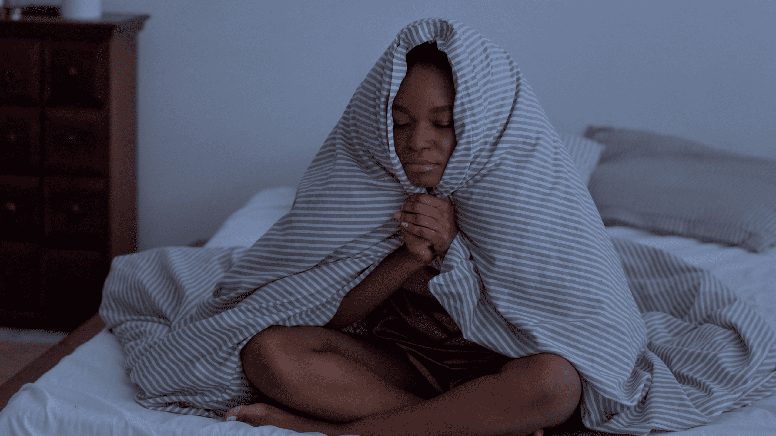 A young girl sitting on her bed at night, wrapped in a striped blanket with her eyes closed, looking restless and unable to sleep. The dim lighting and her expression convey frustration and difficulty winding down.