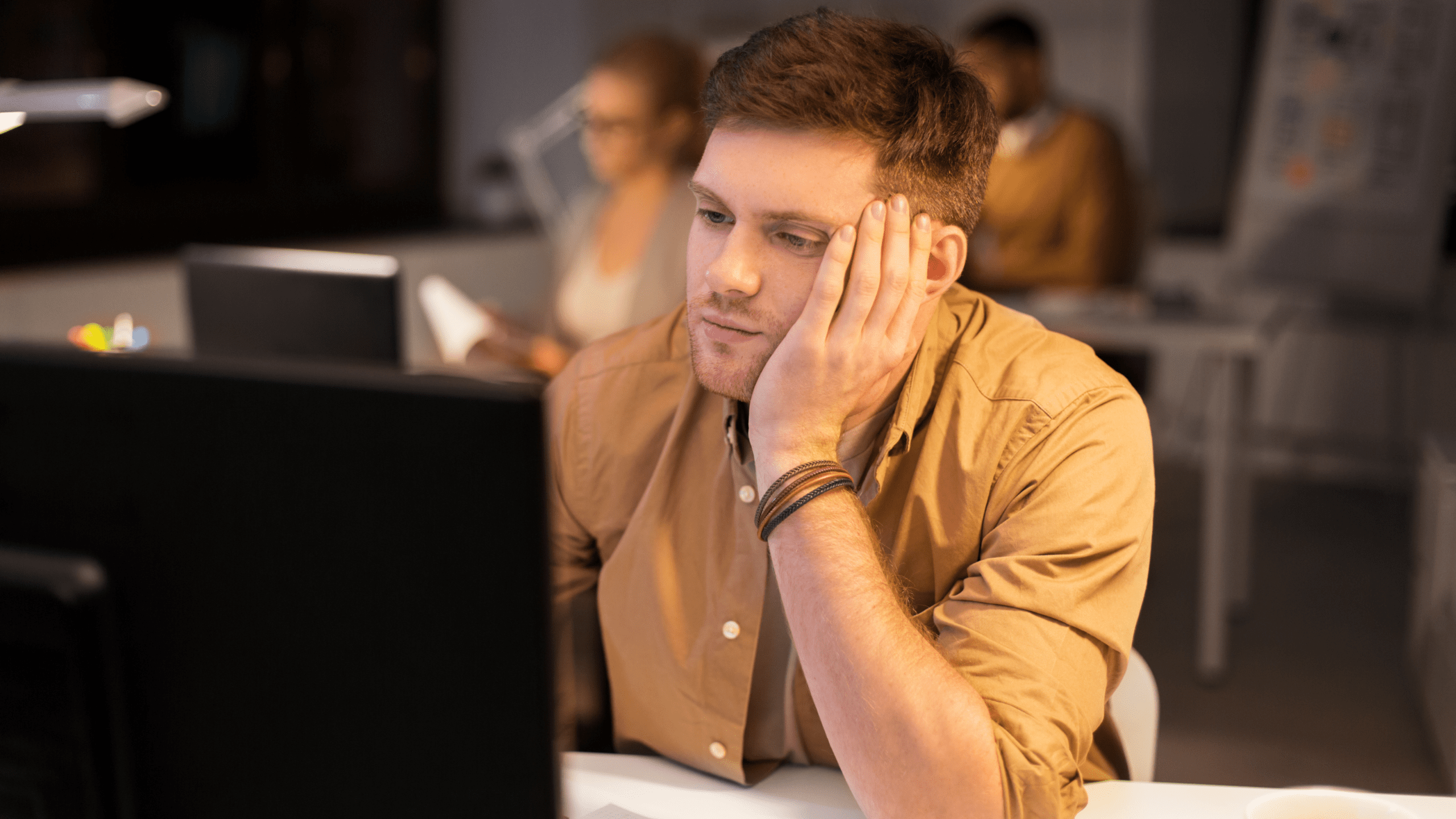 A young man with short brown hair sits at a desk, resting his face on his hand while staring at a computer screen. He appears tired or overwhelmed. The background shows a modern study space with other people working.