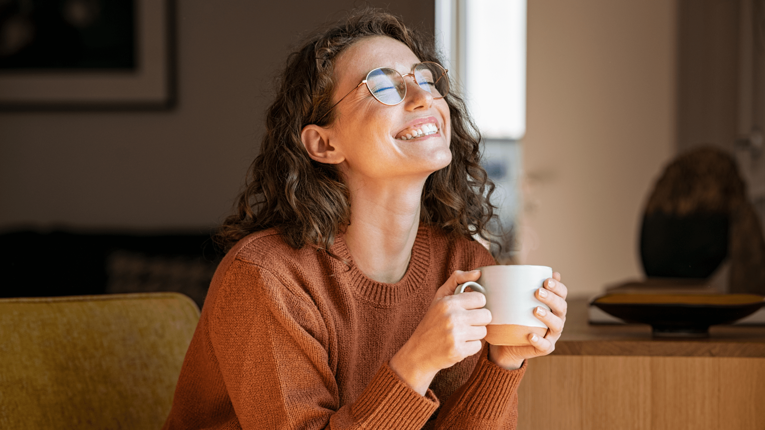 A young woman with curly brown hair and glasses sits on a mustard-yellow chair, smiling with her eyes closed while holding a ceramic mug. She wears a cozy burnt-orange sweater, and soft natural light fills the modern, warmly decorated living space behind her. She looks relaxed and content, savoring a peaceful moment with her drink.